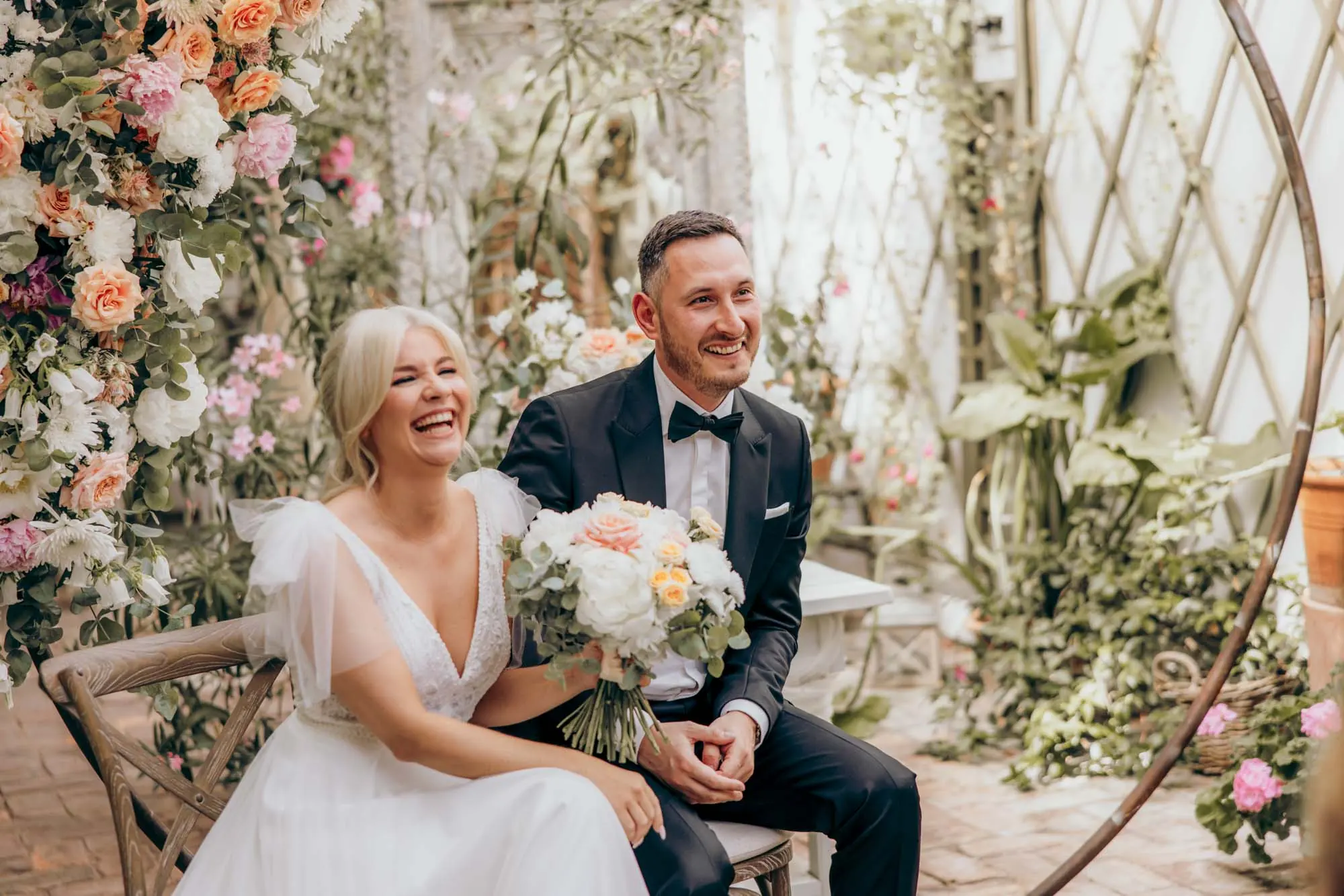 Lachendes Brautpaar mit Brautstrauß zwischen Blumen und Pflanzen in der Alten Gärtnerei München, fotografiert von MOKATI